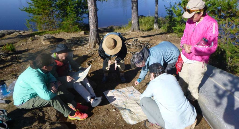 A group of people sit on the shore near a body of water and examine a map that is laid out on the ground. 